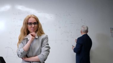 Yaşlı bir adam beyaz tahtaya yazar ve genç bir kadın düşünceli bir şekilde durur.