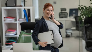 Ofiste cep telefonu kullanan ve kağıt tablet tutan hamile bir kadın.