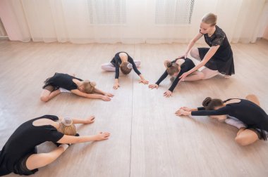 Beyaz kadın ve beş küçük kız bir çember oluşturup bale okulunda esneme hareketleri yapıyorlar.