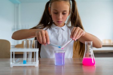 Liseli bir kız kimya dersinde deneyler yapıyor. Beher kabından renkli sıvılar döken bir kız.
