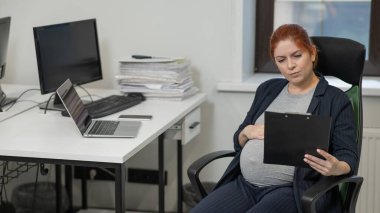 Ofisteki kağıt tablette belgeleri okuyan hamile bir kadın.