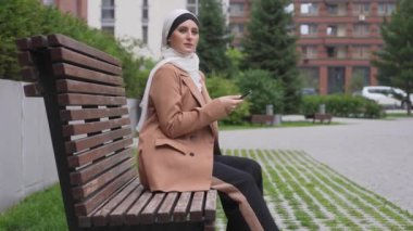 Tesettürlü genç bir kadın açık hava bankında otururken akıllı telefon kullanıyor.