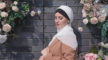 Beyaz bir kadının portresi, çiçekli bir duvarın yanında tesettüre bürünmüş.
