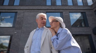 Şapkalı ve gözlüklü bir kadın beyaz ceketli yaşlı bir adama arkadan sarılıyor. Olgun insanların romantik ilişkileri