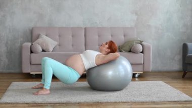 Hamile kızıl saçlı kadın evde fitness topu egzersizleri yapıyor.