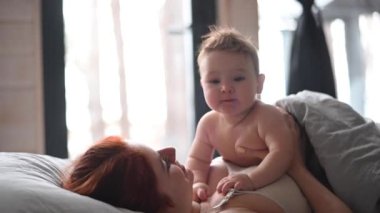 Kızıl saçlı beyaz kadın, panoramik pencereli bir kır evinde bebeğiyle yatakta yatıyor.