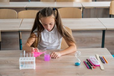 Liseli bir kız kimya dersinde deneyler yapıyor. Beher kabından renkli sıvılar döken bir kız.