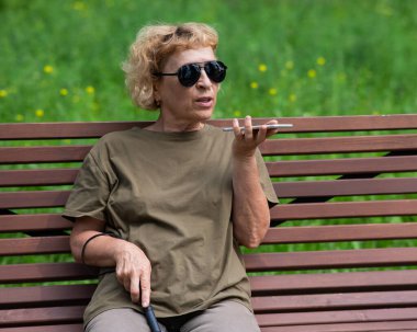 Yaşlı bir kör kadın parkta bir bankta oturur ve akıllı bir telefonla konuşur.