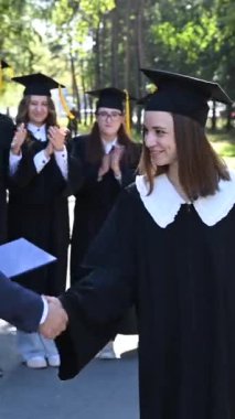 The teacher shakes hands with the student and presents the diploma outdoors. A group of university graduates. Vertical video