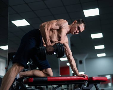 Gömleksiz bir adam spor salonunda dambıl sırasına dizer.