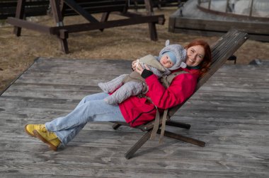 Caucasian woman with her son in an ergo backpack sitting in a wooden deck chair clipart