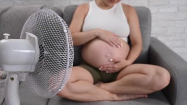 Hamile bir kadın kanepede oturmuş karnını okşuyor ve elektrikli vantilatörün serin havasının tadını çıkarıyor.