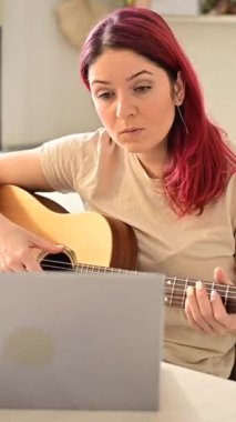 Uzaktan akustik gitar dersi sırasında mutfakta oturan bir kadın. Bir kız gitar çalmayı öğreniyor ve dizüstü bilgisayardan eğitici videolar izliyor. Dikey video