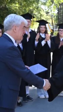 The teacher shakes hands with the student and presents the diploma outdoors. A group of university graduates. Vertical video