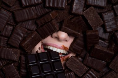 The face of a caucasian woman surrounded by chocolates. The girl eats a bar of chocolate