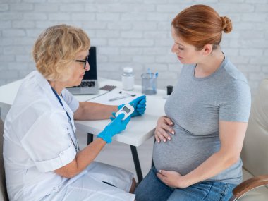 Yaşlı bir kadın doktor hamile kızıl saçlı bir kadının kan şekerini ölçüyor. Gebelik Diyabet