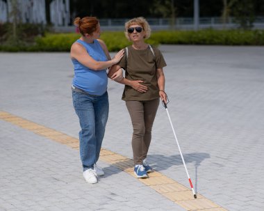 Caucasian pregnant woman leading blind elderly lady outdoors clipart