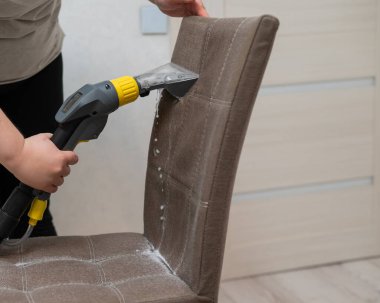 Kadın, elektrikli süpürgeyle kumaş sandalyesinin arkasını temizliyor.