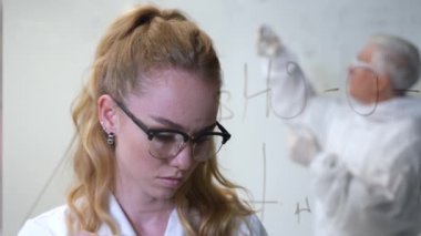 Kadın bir kimyager cama bir formül yazar. Koruyucu giysili yaşlı bir beyaz adam testler yapıyor.