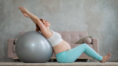 Hamile kızıl saçlı kadın evde fitness topu egzersizleri yapıyor.