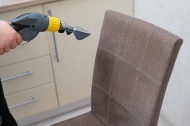 Kadın, elektrikli süpürgeyle kumaş sandalyesinin arkasını temizliyor.
