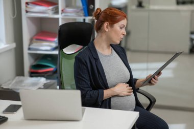 Ofisteki kağıt tablette belgeleri okuyan hamile bir kadın.