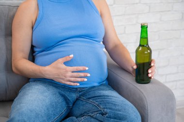 Suratsız hamile kadın kanepede otururken bira içiyor.