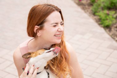 Köpek Jack Russell Terrier sahibinin yüzünü açık havada yalıyor. Dişlerinde diş teli olan kız.