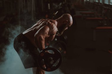 Beyaz, kel, üstsüz bir adam spor salonunda halterle egzersiz yapıyor.