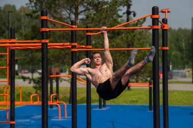 Genç kaslı bir adam tek eliyle açık havada yatay bir barda asılı duruyor.