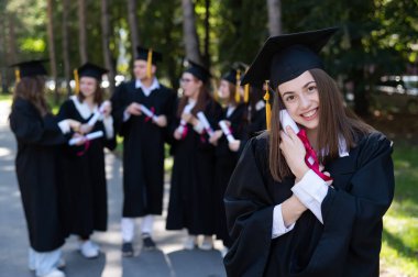 Group of happy students in graduation gowns outdoors. A young girl boasts of her diploma