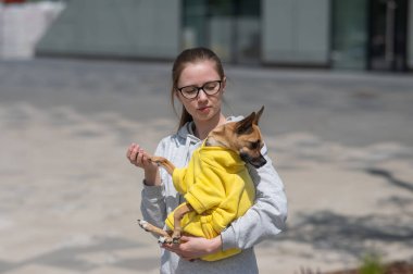 Kafkasyalı genç bir kadın sarı tulumlu küçük bir köpeği elinde tutuyor.
