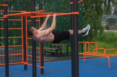 Üstsüz erkek sporcu ön tarafta yatay barda asılı duruyor.