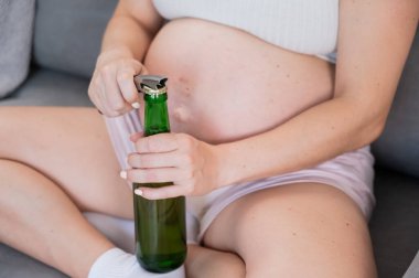 Kanepede oturan ve bira şişesini açan yüzü olmayan hamile kadın.