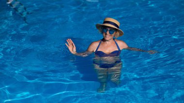 Havuzda mavi gözlüklü ve şapkalı yaşlı bir kadının portresi yüzüyor.