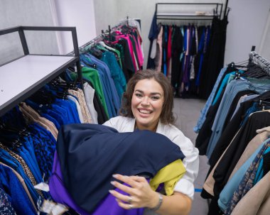 Şişko yanaklı bir kadının kucak dolusu giysiyi büyük boy bir mağazada tutuşu.