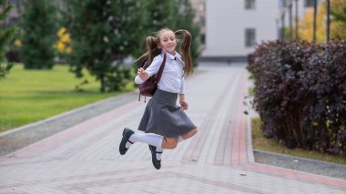 Üniformalı ve sırt çantalı mutlu beyaz kız okuldan sonra dışarı atlıyor.