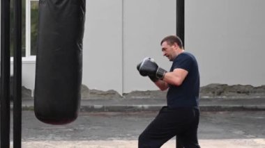 Orta yaşlı bir adam dışarıda kum torbasıyla boks yapıyor.