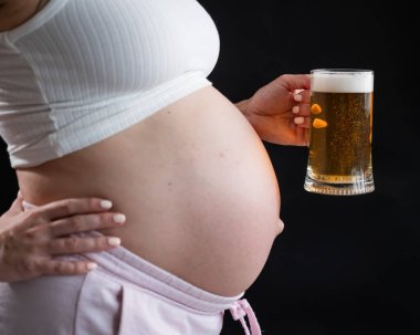 Siyah arka planda elinde bir bardak bira tutan hamile bir kadının göbeğine yakın çekim. Deri döküntüsü