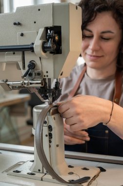 A woman tanner sews a leather belt on a sewing machine. Vertical photo clipart