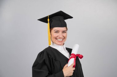 Mezuniyet elbisesi içinde gülümseyen kadın beyaz arka planda diploma taşıyor.