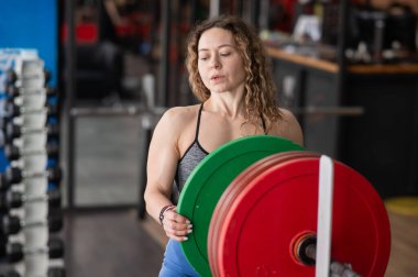 Kırk yaşında beyaz bir kadın spor salonundaki bir halterin üzerine ağırlık plakası koyuyor.