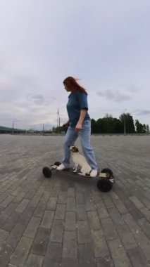 Beyaz kadın elektrikli uzun tahta üzerinde Jack Russell Terrier köpeği ile geziyor. Dikey video