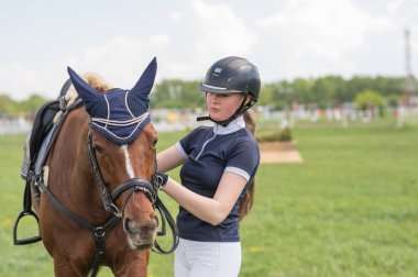 Genç bir kız bir binicilik yarışmasının önünde bir atın yanında duruyor.