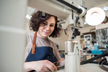 A woman tanner sews a leather belt on a sewing machine clipart