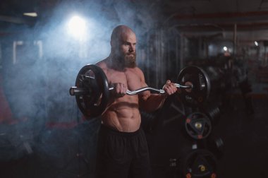 Beyaz, kel, üstsüz bir adam spor salonunda halterle egzersiz yapıyor. Ağırlıklarla bibik bukleler