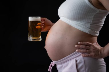 Siyah arka planda elinde bir bardak bira tutan hamile bir kadının göbeğine yakın çekim. Deri döküntüsü