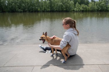 Genç beyaz bir kadın ve şirin küçük bir köpek nehir kıyısında uyumlu spor ayakkabılarla yürüyorlar.