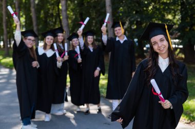 Mezuniyet elbisesi giymiş bir grup mutlu öğrenci dışarıda. Ön planda elinde diploması olan genç bir kız.