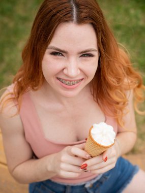 Diş telleriyle gülümseyen ve açık havada dondurma yiyen güzel kızıl saçlı bir kadın. Dikey fotoğraf